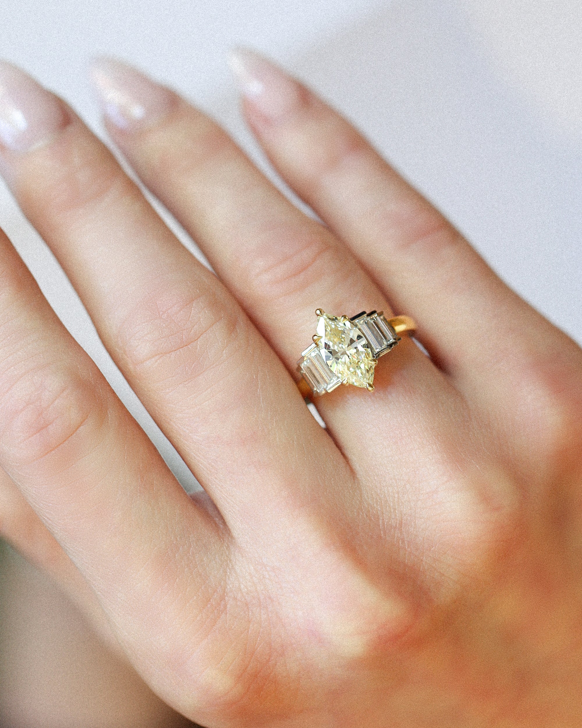 A close-up of a hand wearing a yellow gold ring with a central marquise-cut yellow diamond and baguette side stones, set against a soft light gray background.