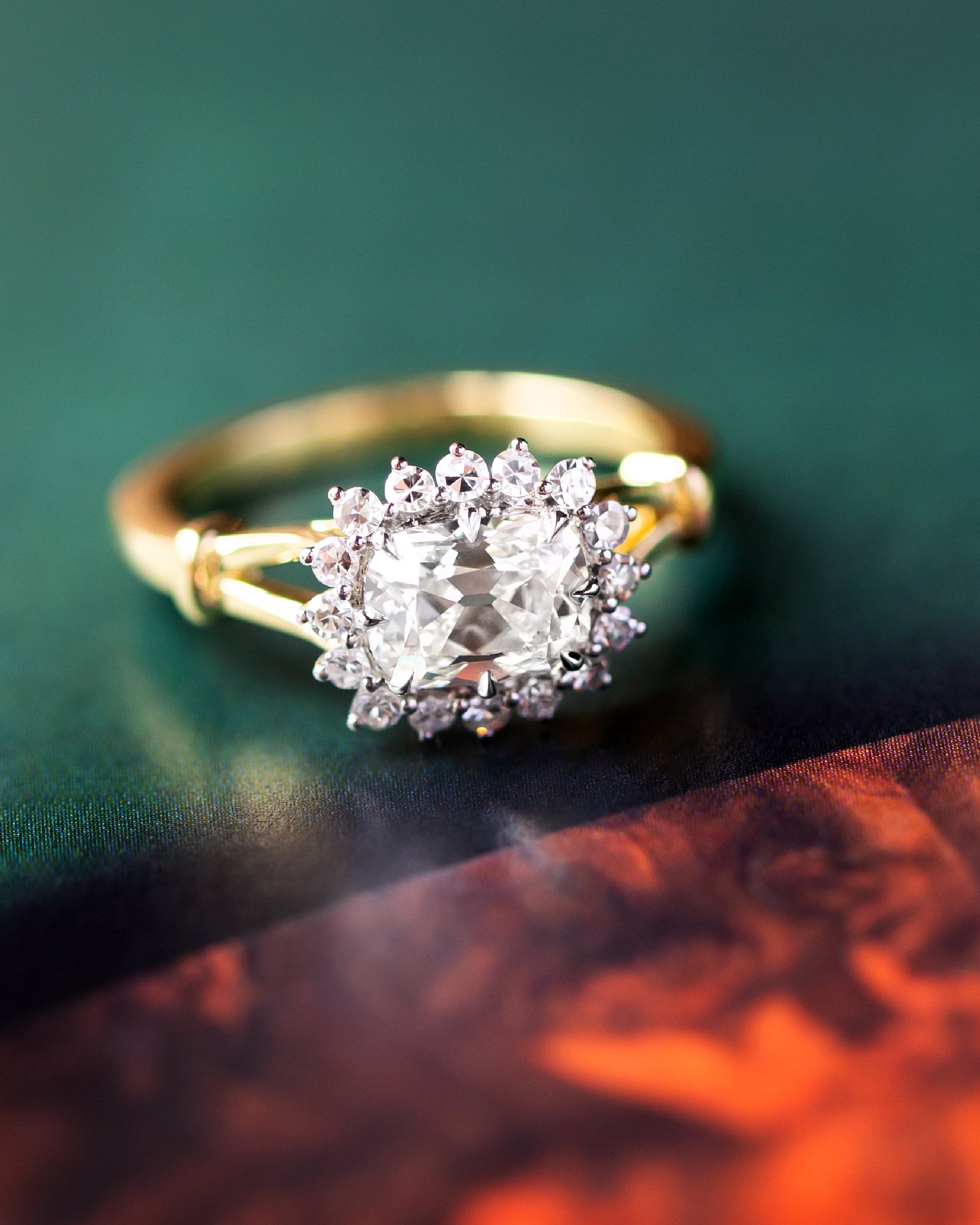 A yellow gold ring with a cushion-cut diamond center stone and a halo of small round diamonds, photographed close up on a dark green surface with a blurred orange-toned area in the foreground.