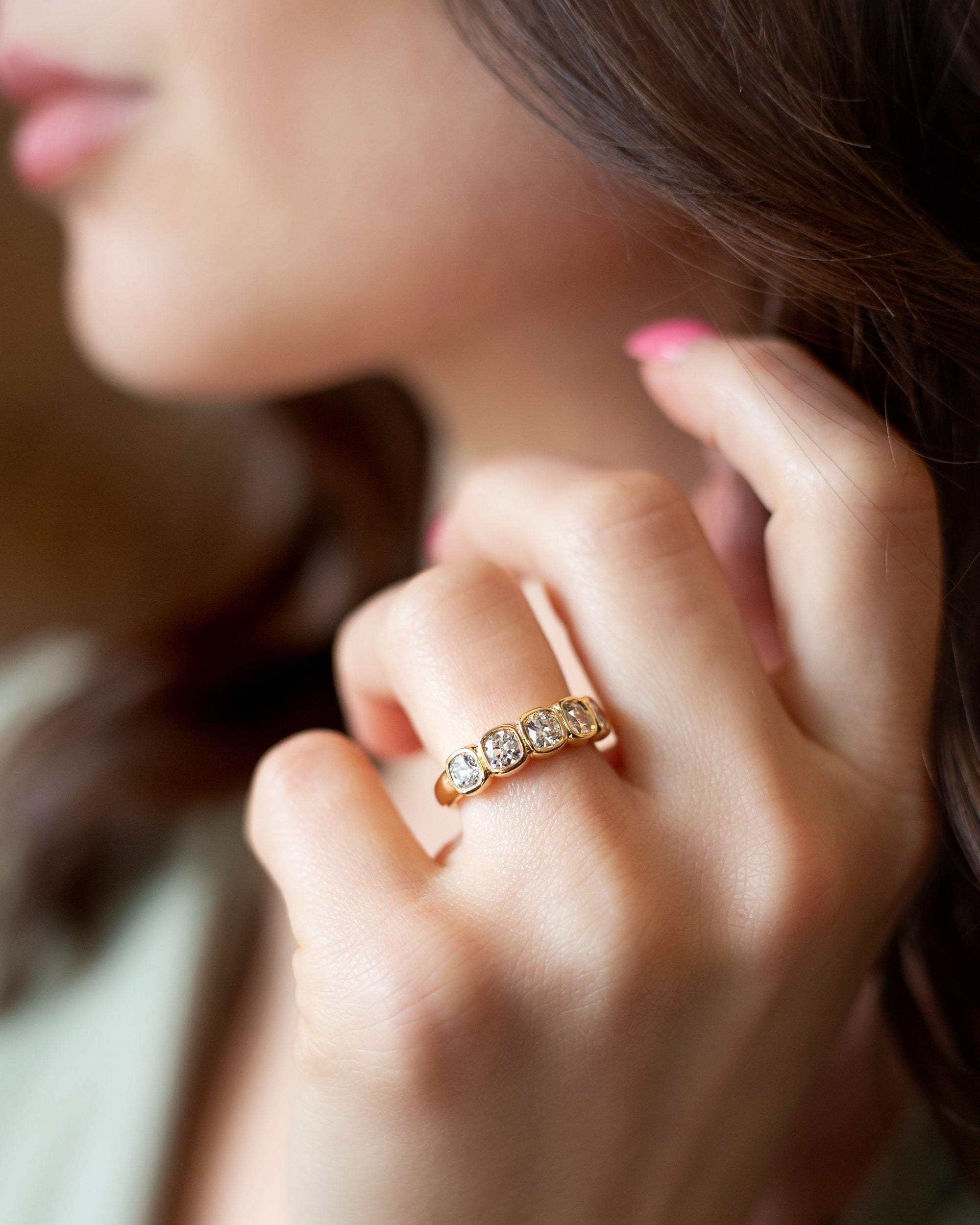 A close-up lifestyle shot of a woman's hand wearing a gold diamond band ring with five Old Mine cut diamonds, bezel-set against a soft-focus background of her face and hair.