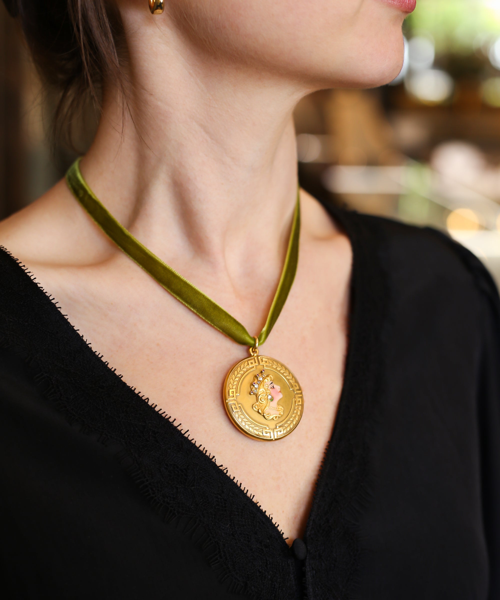 A close-up lifestyle shot of a woman wearing an Art Nouveau gold pendant necklace with an olive green velvet ribbon. The pendant features a gold portrait of woman in profile, wearing a crown and gem-set earring.