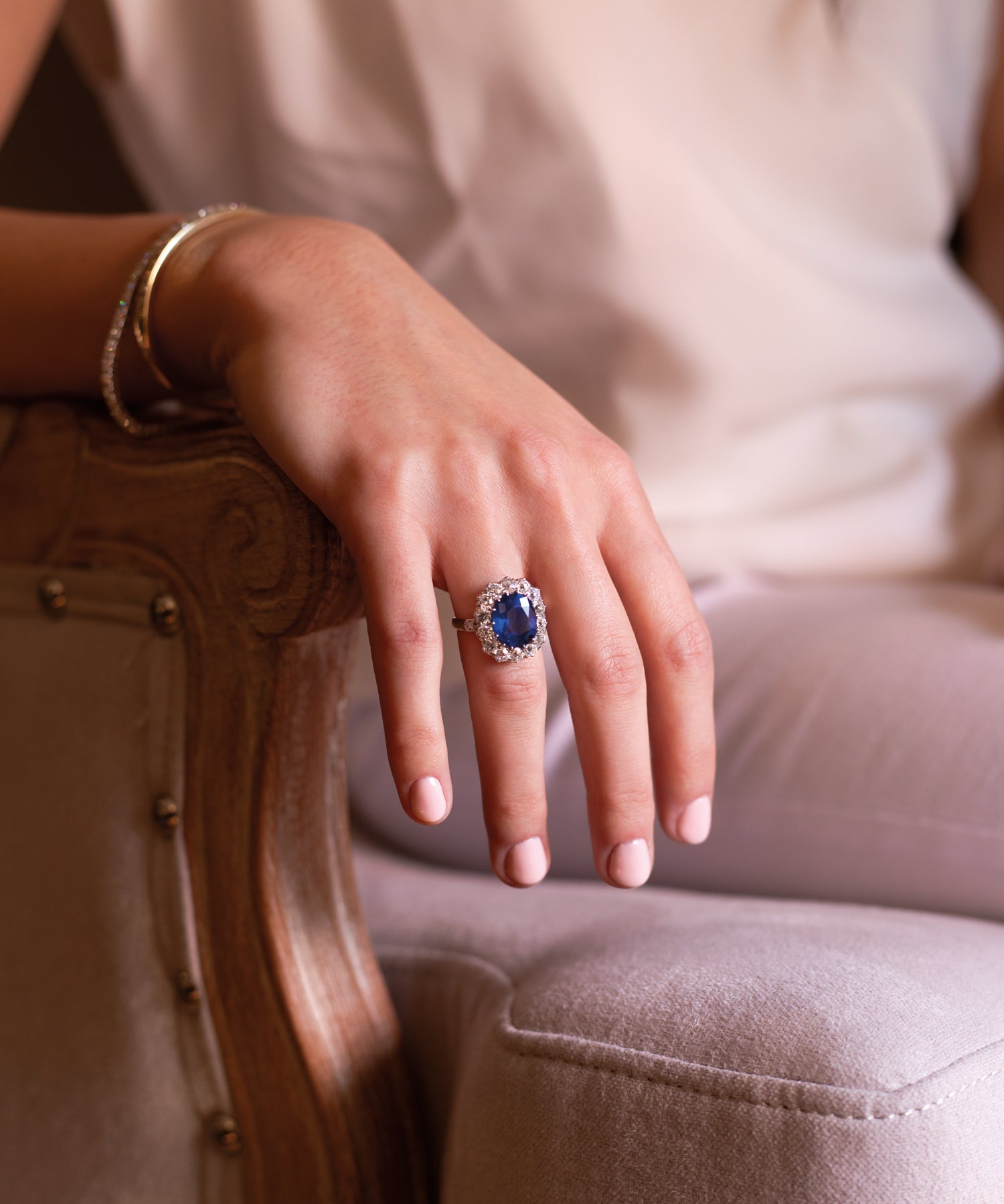 Close-up of a hand wearing a ring with a blue sapphire and diamonds, sitting on a textured chair.