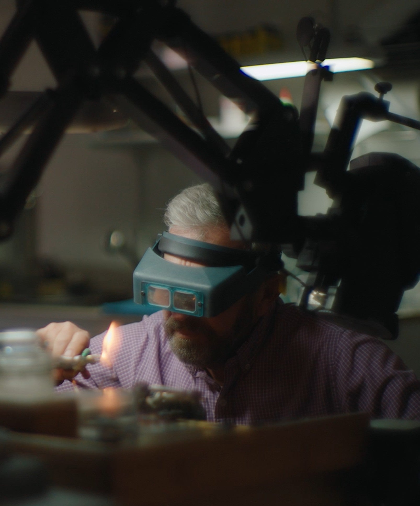 A man wearing a teal jeweler's loupe magnifier and a purple checkered shirt, holding a flame in a workshop under a large black mechanical arm.